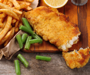 Breaded fried fish fillet and potatoes with asparagus and lemon on cutting board and rustic wooden background